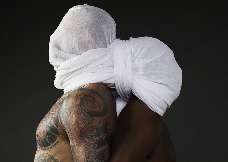 two men stand back to back with a white sheet twisted over their heads. the man on teh left is taller and heavily tattoed, the an on the right is shorter with a darker complexion.