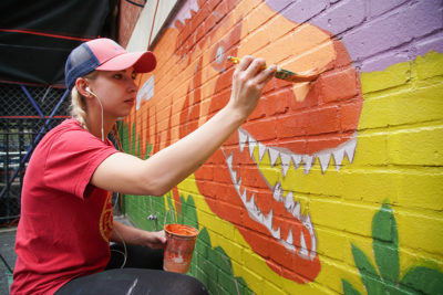 blonde woman in ball cap painting dinosaur on brick wall