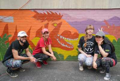 one man, two woman, crouch in front of mural of dinosuar