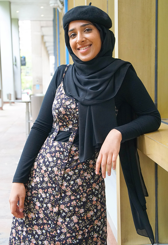 smiling young woman in black beret and black headscarf with black long sleeve shirt under floral print dress posing outside