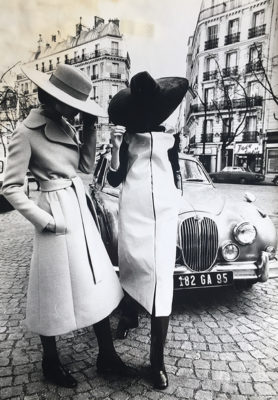 Two models posing on the streets of Paris, in large brimmed suede and felt hats, paired with a belted, wool, knee length coat and a vinyl, knee length, raincoat, over dark colored pants and shoes