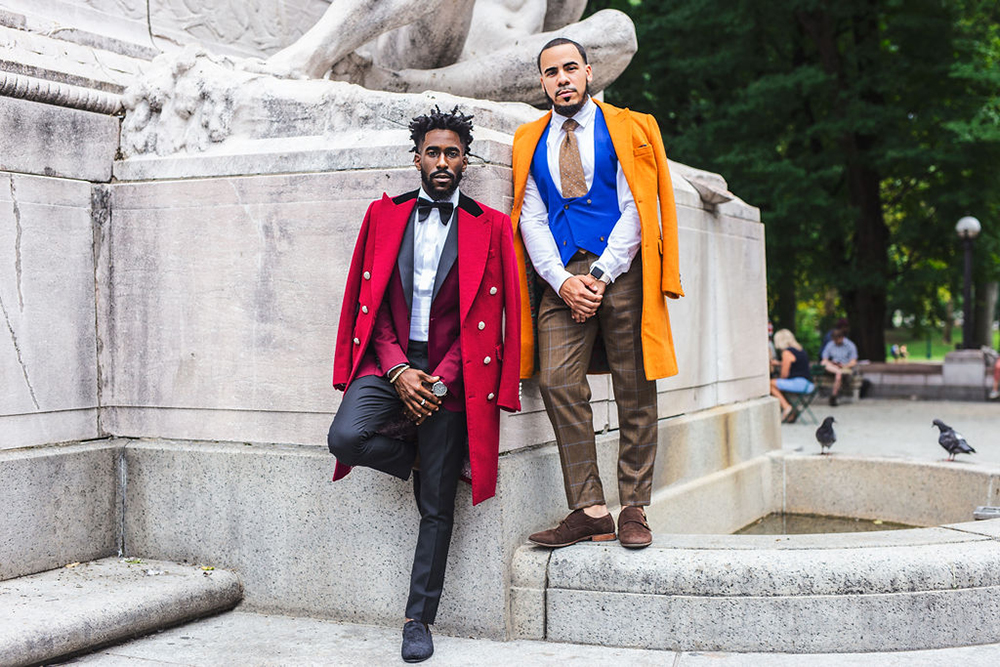 Two men in suits with overcoats. Left, a red peak lapel cashmere overcoat and tuxedo. Right, a pumpkin spice wool overcoat, blue vest, and brown windowpane pants.