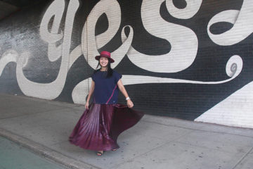 woman in navy top, burgundy ankle length skirt and burgundy hat in front of a mural reading "yes!"