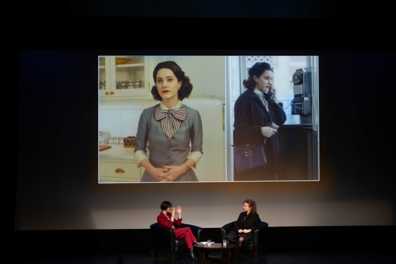 two women sitting on stage under a screen with a scene from the TV show The Marvelous Mrs. Maisel