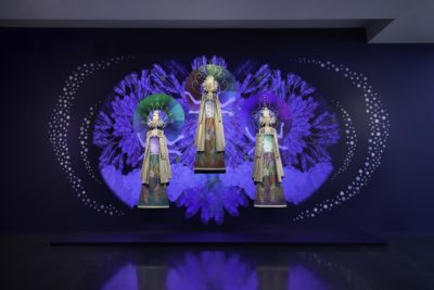 Image of an art installation by artist Saya Woolfalk, featuring three figures against a deep blue background.
