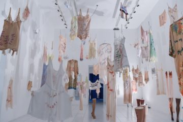 View of an art installation with many garments hanging from the ceiling in a gallery.