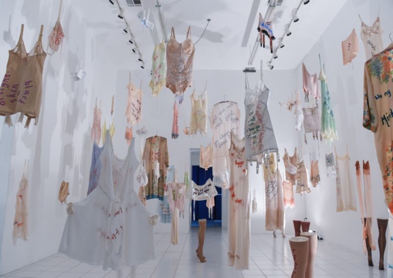 View of an art installation with many garments hanging from the ceiling in a gallery.