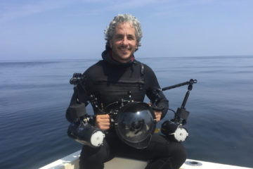 Professor in wetsuit holding underwater camera with ocean in background