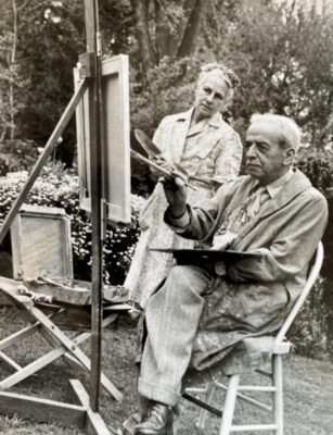 Black and white photo of Max Meyer sitting outside at an easel with his wife standing and watching