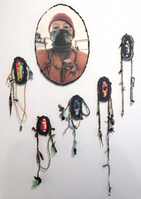 oval image of a masked woman with five artifacts encircling the bottom that have woven strands that dangle from each