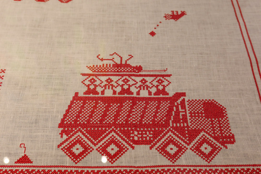 red embroidery on a natural linen background of a tank with figures holding a cockroach above them