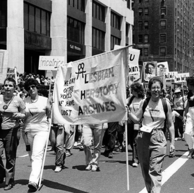 Black and white vintage photo of a march with women holding a sign that says Lesbian Herstory Archive