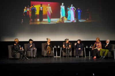 several panelists sitting onstage in chairs with footage of Stephen Burrows' clothes behind them