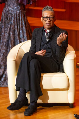 B Michael in a suit gesticulating in a white armchair with a flowy purple-gray dress behind him