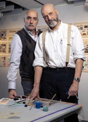 Two well-dressed men standing close with paints on a table in front of them