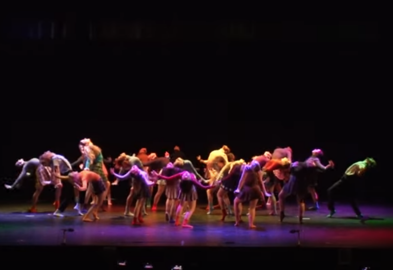 dozens of dancers doused in colorful light all leaning back away from each other