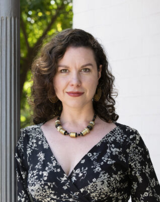 Hilary Davidson headshot. A woman with shoulder-length brown hair wears a beaded necklace and a black V-neck wrap top with a white floral print. She leans against a concrete gray pillar.