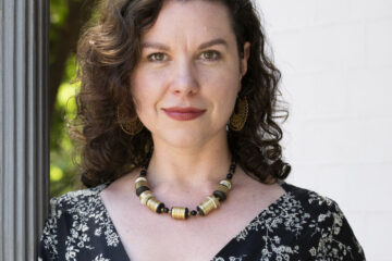 Hilary Davidson headshot. A woman with shoulder-length brown hair wears a beaded necklace and a black V-neck wrap top with a white floral print. She leans against a concrete gray pillar.