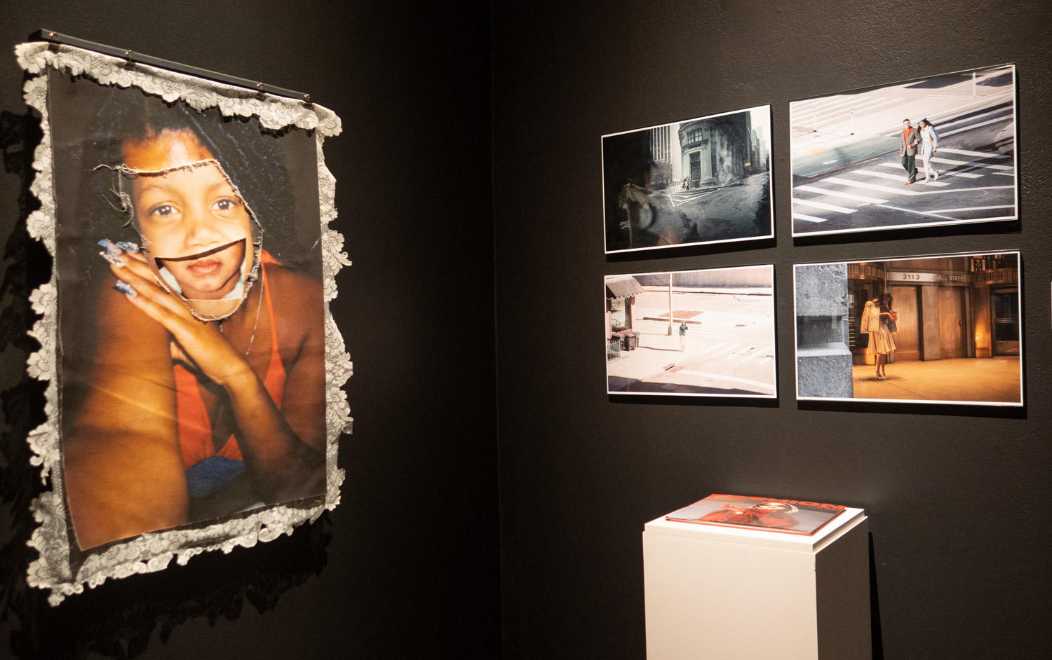 Photographs on a dark wall, on of a Black woman with a girl's mouth, four more of people in cities