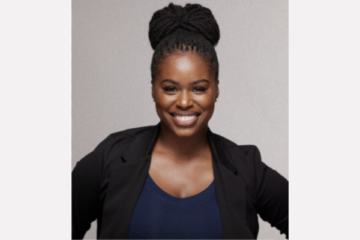Co-director of FIT Dance Company Quiantae Mapenzi Johnson smiles at the camera. She has her black hair in a topknot and wears a black open blazer with a navy blue scoop-neck top underneath.