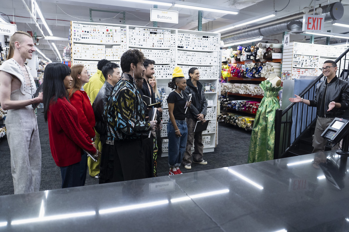 a group of people stand in Mood Fabrics store listening to a person