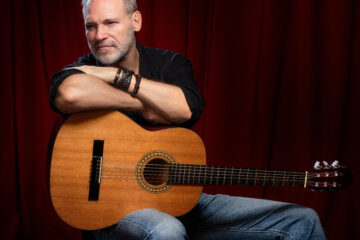 Actor Jim Newman sits with an acoustic guitar in a photographic portrait shot by FIT Professor Ron Amato.