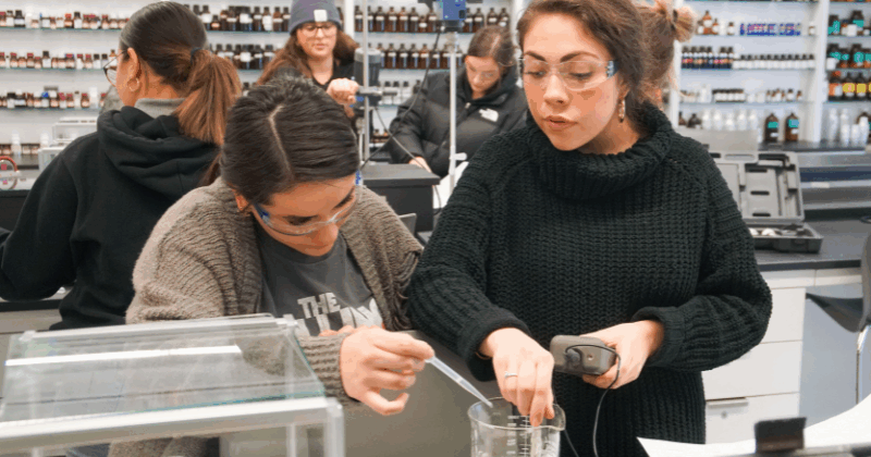 several students perform experiments in the FIT fragrance lab, two in the foreground are working with a beaker filled with white fluid
