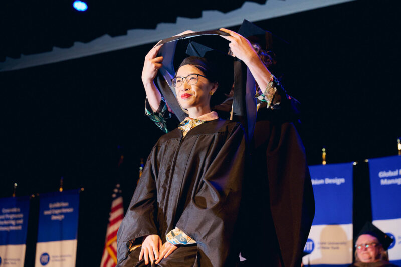 A student in a black commencement gown receives a hood atop her head