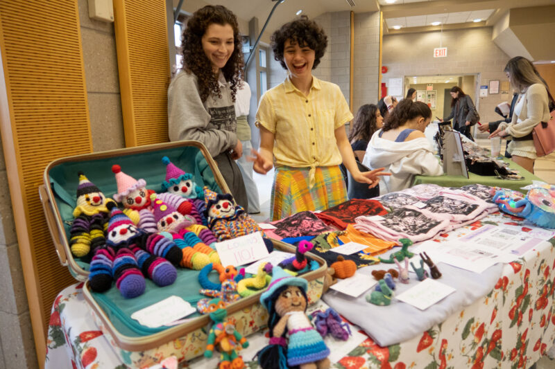 Two smiling students selling knitted dolls at a table indoors