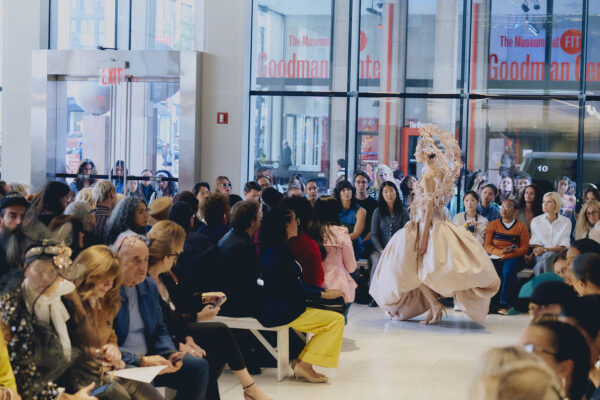 Model in an elaborate white dress walking toward an audience in a glass-walled room