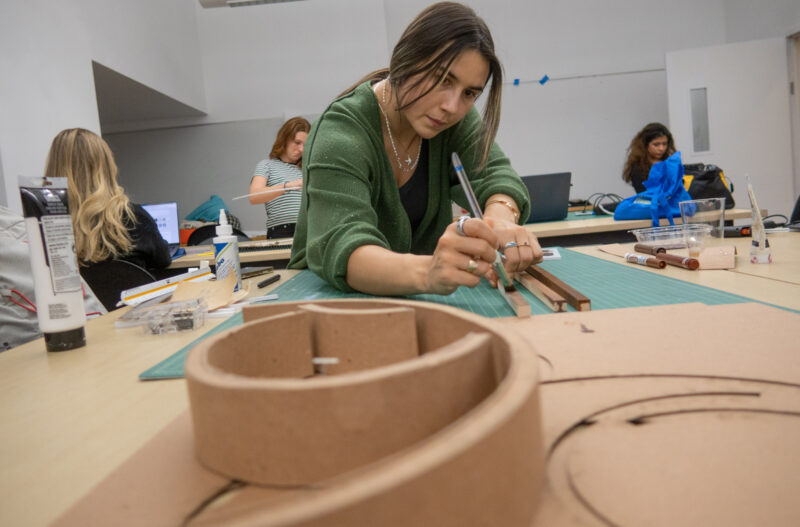 A student paints strips of wood; a curved scale model is in the foreground