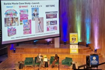 Two panelists sitting in a wood-paneled auditorium with a slide about Barbie on a large projection screen