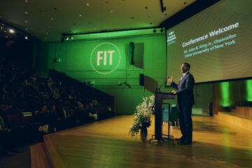 Pulled-out shot of man in suit standing at podium with FIT logo in the background