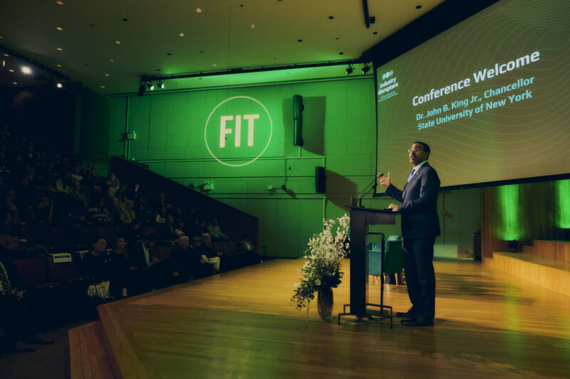 Pulled-out shot of man in suit standing at podium with FIT logo in the background