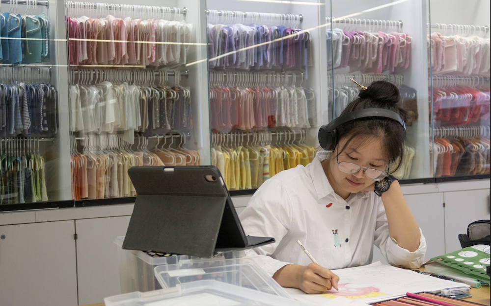 student with headphones sitting at a table with a tablet computer and shelves of colorful fabric samples behind her behind glass