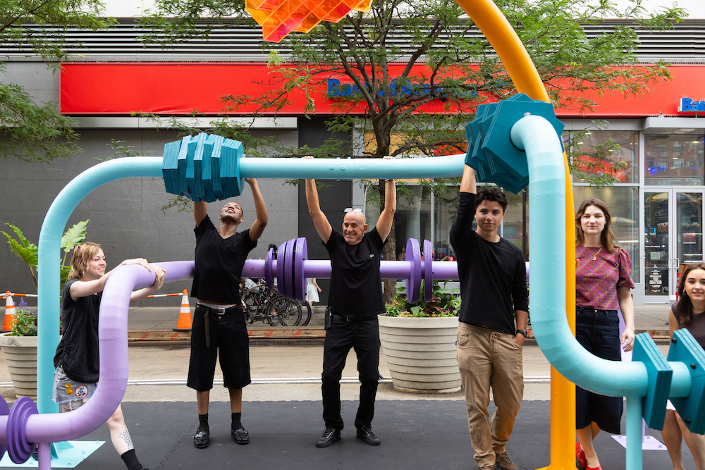 people touch and hang on large colorful pipes that are part of an outdoor public sculpture