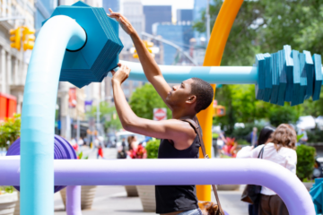 student manipulates a large blue plate on a metal tube of a public sculpture; other colored tubes of the sculpture surround him