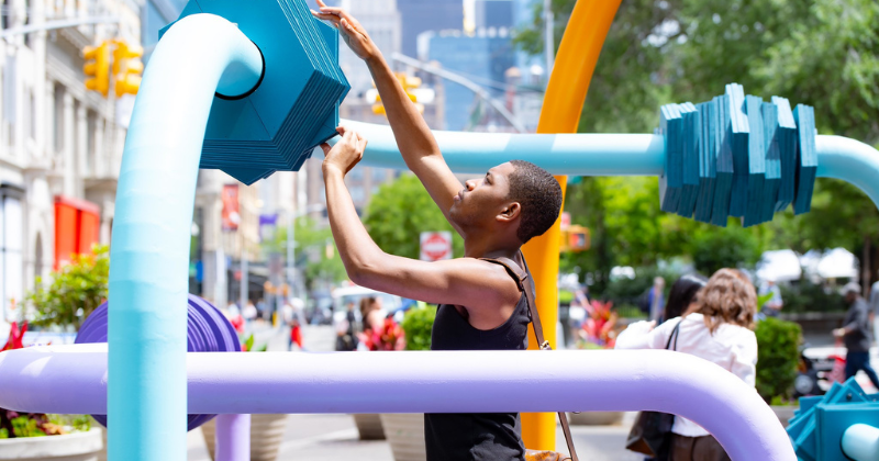 student manipulates a large blue plate on a metal tube of a public sculpture; other colored tubes of the sculpture surround him
