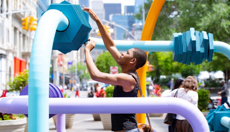 student manipulates a large blue plate on a metal tube of a public sculpture; other colored tubes of the sculpture surround him