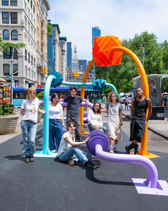 Seven students stand amongst the multicolored pipes that comprise the public sculpture they designed that on display on a New York pedestrian island