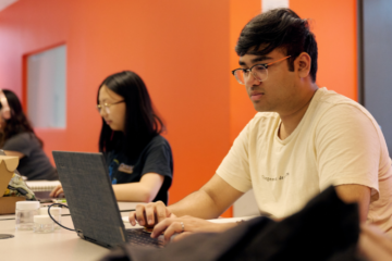 three students sit at laptops in a computer lab with orange wall behind them