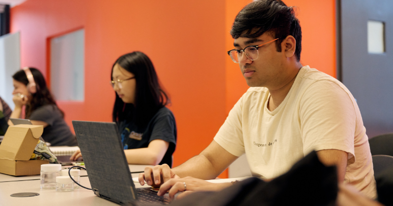 three students sit at laptops in a computer lab with orange wall behind them
