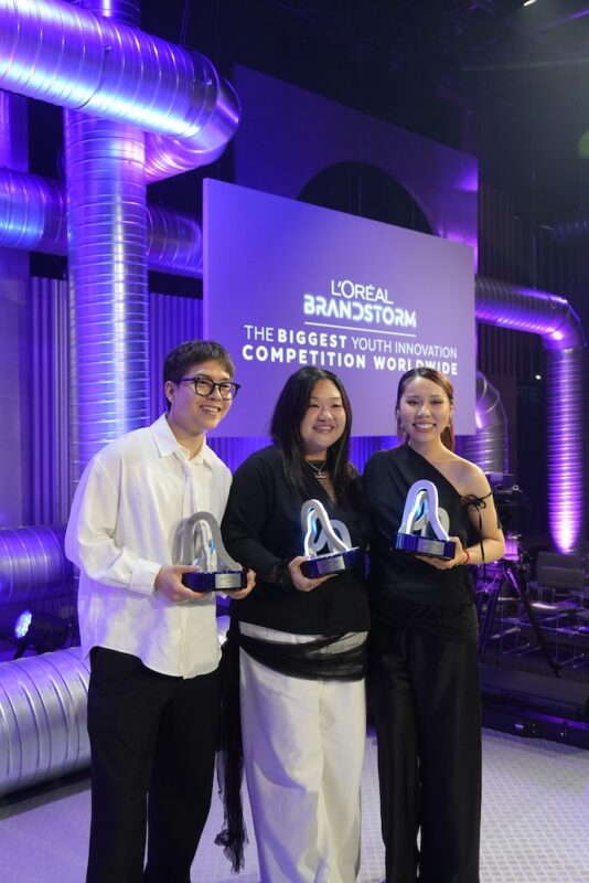 three smiling students stand holding their trophies on a purple lit stage