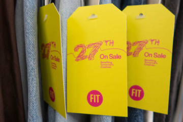 A row of hanging clothes with large yellow hangtags with text 27th on Sale Benefiting Tomorrow's Creatives