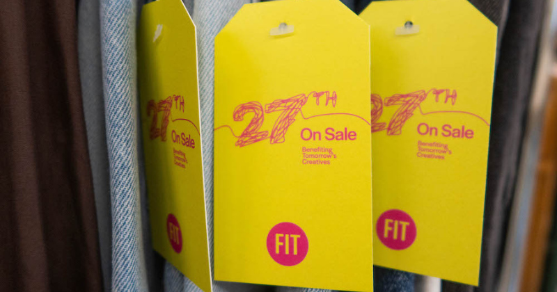 A row of hanging clothes with large yellow hangtags with text 27th on Sale Benefiting Tomorrow's Creatives